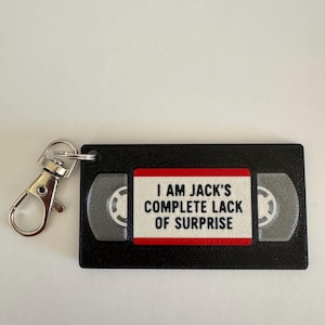 May include: A black rectangular keychain shaped like a VHS tape with a silver metal lobster clasp. The center label is white with a red border and features the black text: I AM JACK'S COMPLETE LACK OF SURPRISE. The tape reels are grey.