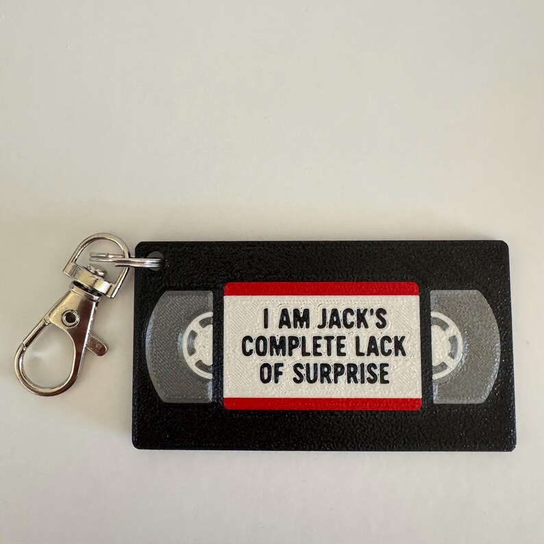 May include: A black rectangular keychain shaped like a VHS tape with a silver metal lobster clasp. The center label is white with a red border and features the black text: I AM JACK'S COMPLETE LACK OF SURPRISE. The tape reels are grey.