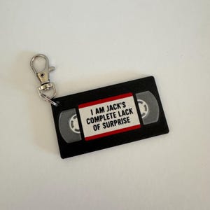 May include: A black keychain shaped like a VHS tape with a white label featuring the red-bordered text I AM JACK'S COMPLETE LACK OF SURPRISE. The rectangular charm includes a silver-toned metal lobster clasp attached to the top left corner.
