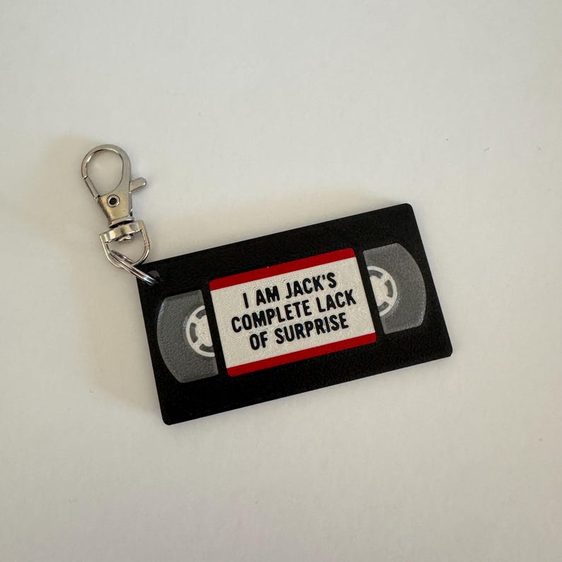May include: A black keychain shaped like a VHS tape with a white label featuring the red-bordered text I AM JACK'S COMPLETE LACK OF SURPRISE. The rectangular charm includes a silver-toned metal lobster clasp attached to the top left corner.