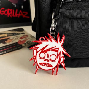 May include: A white and red acrylic keychain featuring a stylized cartoon character head with spiky hair, wide circular eyes, and an open mouth. The keychain is attached to a silver-toned metal lobster clasp hook hanging from a black fabric bag.