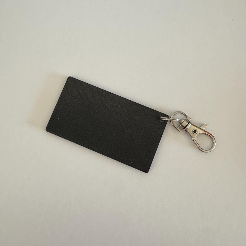May include: A rectangular black keychain featuring a textured surface and a silver-toned metal lobster clasp attached to one corner by a small jump ring. The object is flat and solid, presenting a simple design with a functional metal hardware component.