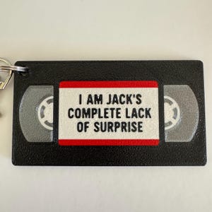 May include: A black rectangular keychain shaped like a VHS tape with a white center label featuring the black text I AM JACK'S COMPLETE LACK OF SURPRISE. The design includes a red border around the label and two grey circular reel details.