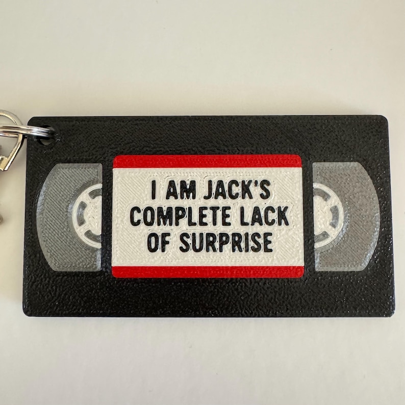 May include: A black rectangular keychain shaped like a VHS tape with a white center label featuring the black text I AM JACK'S COMPLETE LACK OF SURPRISE. The design includes a red border around the label and two grey circular reel details.