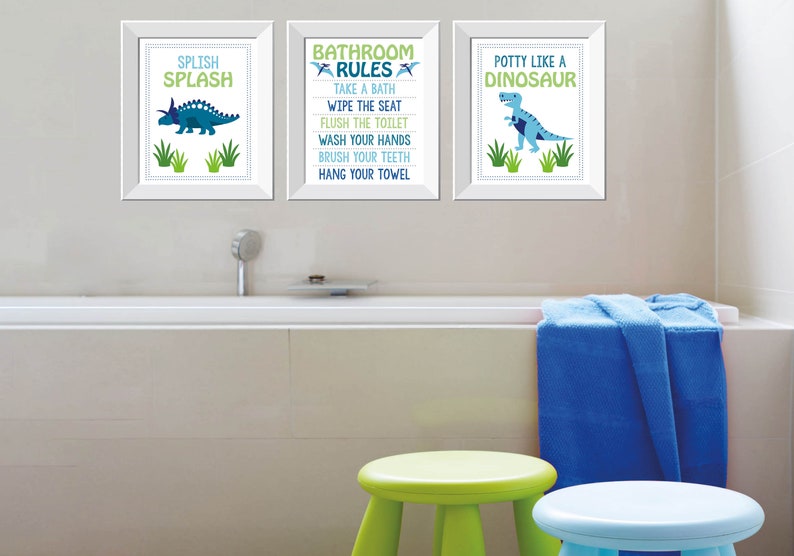 May include: Three framed prints with dinosaur illustrations and text. The prints say "Splish Splash", "Bathroom Rules", and "Potty Like a Dinosaur". The color scheme is blue, green, and white. The scene includes a bathtub, stools, and a blue towel.