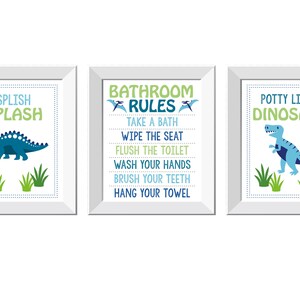 May include: Three framed prints with dinosaur-themed bathroom decor. The prints feature text like "Splish Splash," "Bathroom Rules," and "Potty Like a Dinosaur," along with dinosaur illustrations in blue and green. The frames are white.