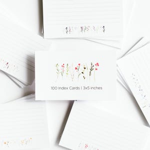 Könnte beinhalten: Weiße Karteikarten mit linierten Seiten und einem floralen Design. Die Karten sind 7,6 x 12,7 cm groß und zeigen eine Aquarellillustration von Wildblumen. Der Text auf der Karte lautet "100 Index Cards | 3x5 inches".