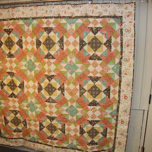 Può includere: Un grande quilt con un motivo geometrico in tonalità di marrone, verde, rosa e blu. Il quilt è composto da quadrati e triangoli, e il motivo si ripete su tutto il quilt.