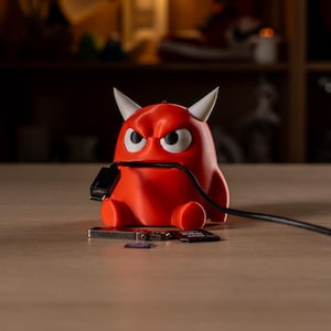 May include: A red, horned, monster-shaped cable organizer figurine with white eyes and a furrowed brow. The figure holds a black USB cable in its mouth, with a silver USB flash drive and a black SD memory card resting on the surface in front of it.