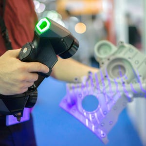 May include: A handheld black 3D laser scanner with a glowing green hexagonal light indicator, projecting a series of parallel purple laser lines onto a complex grey metallic mechanical engine component to capture its precise surface geometry.