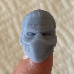 May include: A small, gray, 3D-printed miniature head sculpture featuring a stylized mask that covers the nose and mouth area, with defined brow ridges, recessed eye sockets, and two small circular indentations on each side of the mask.