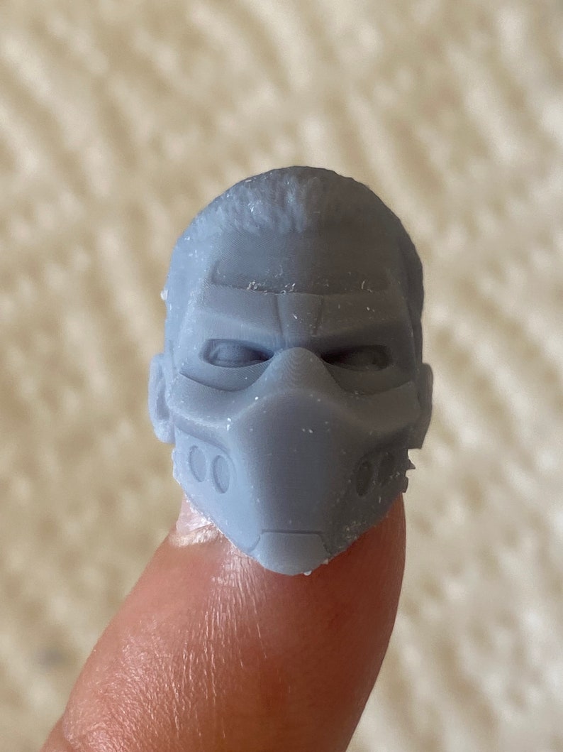 May include: A small, gray, 3D-printed miniature head sculpture featuring a stylized mask that covers the nose and mouth area, with defined brow ridges, recessed eye sockets, and two small circular indentations on each side of the mask.