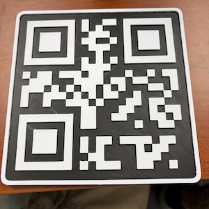 May include: A square, flat, black and white 3D-printed QR code sign with a raised white border and white pixelated pattern on a black background. The design features a standard square matrix layout with three large corner squares and a central grid.
