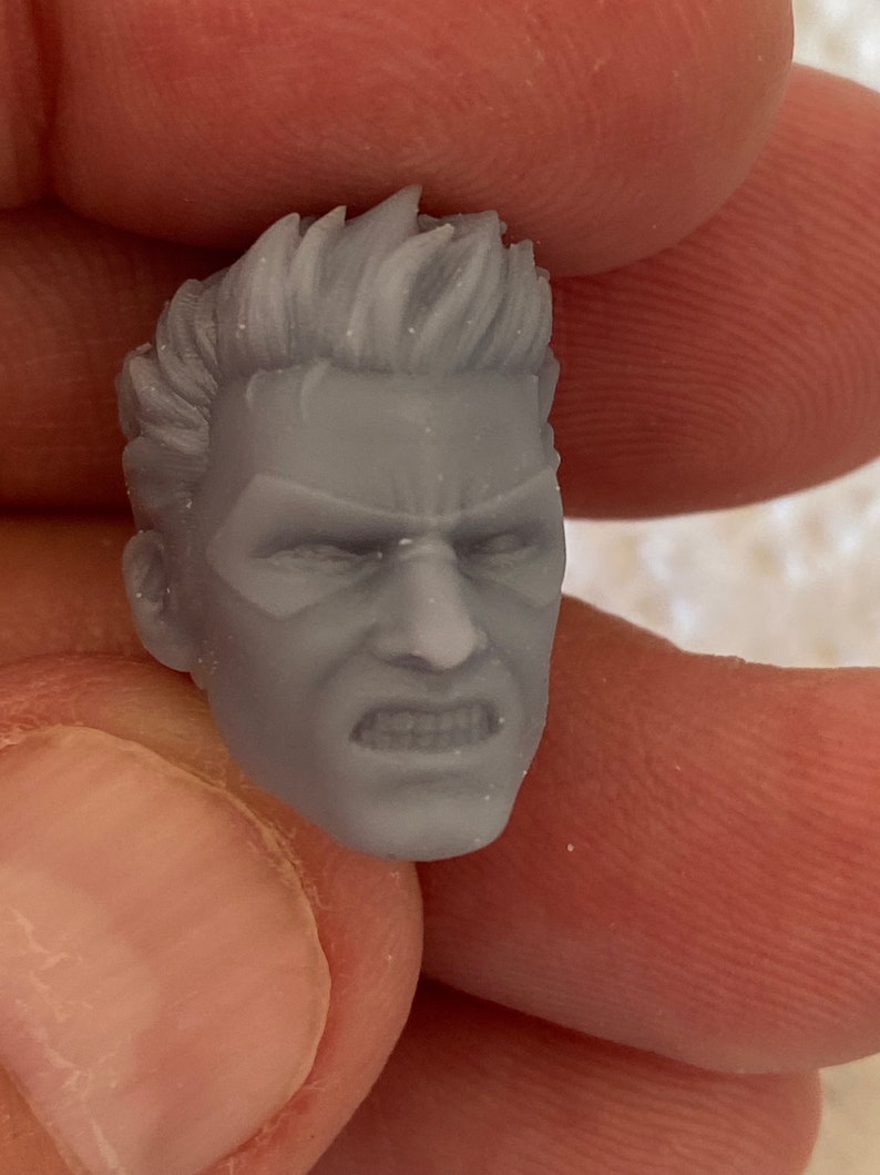 May include: A small, unpainted gray resin miniature head featuring a character with short, spiked hair, a furrowed brow, and a clenched, gritted-teeth expression. The facial features include defined cheekbones and a sharp jawline, sculpted with fine detail.