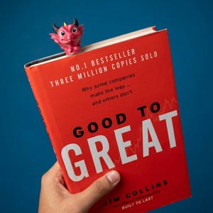 May include: A red dragon-shaped bookmark with black horns and a yellow tip, perched on the top edge of a red book titled Good to Great by Jim Collins, which features white and black text on its cover against a solid blue background.