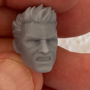 May include: A small, unpainted gray resin miniature head featuring a character with short, spiked hair, a furrowed brow, and a clenched, gritted-teeth expression. The facial features include defined cheekbones and a sharp jawline, sculpted with fine detail.