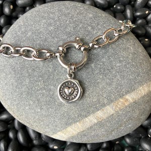 May include: A silver chain bracelet with a heart-shaped charm. The charm is attached to a ring that is part of the chain. The bracelet is on a gray stone background.