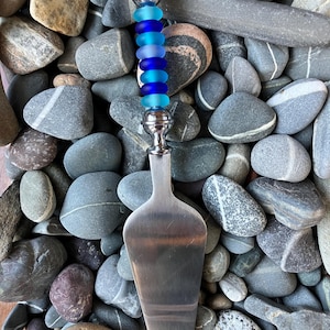 May include: A silver cake server with a blue glass bead handle. The handle is made of multiple beads in shades of blue, from light to dark. The server is resting on a bed of gray and white stones.
