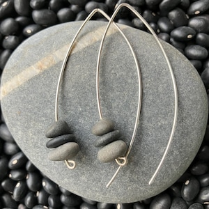 May include: Silver-toned, curved earrings with small, stacked gray stones. The earrings are displayed on a gray stone, set against a backdrop of black beans. The design is minimalist and natural, suitable for everyday wear.