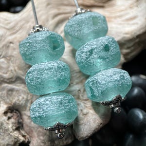 May include: A pair of earrings featuring three stacked, frosted, seafoam green glass beads on each silver-toned metal pin. The beads have a weathered, textured surface and are accented with small, decorative silver-toned metal caps at the top and bottom.
