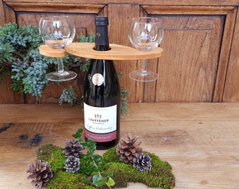 May include: Wooden wine bottle holder with two glass wine glasses attached. The holder is on a wooden surface with moss and pine cones.