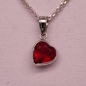 Puede incluir: Un collar de plata con un colgante en forma de corazón. El colgante presenta una piedra preciosa roja oscura facetada engastada en un bisel de plata. La cadena es un diseño delicado y enlazado. El fondo es de color rosa claro.