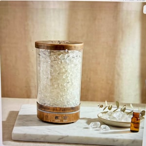 May include: A cylindrical essential oil diffuser with a wooden base and top, and a clear glass container filled with white stones. The base has light and mist control buttons. A small amber bottle and a dish with stones are next to the diffuser.
