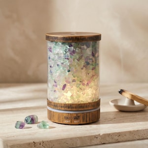 May include: A cylindrical essential oil diffuser with a wooden base and top. The clear glass container is filled with colorful, polished stones. The base has light and mist control buttons. A small dish and incense are in the background.