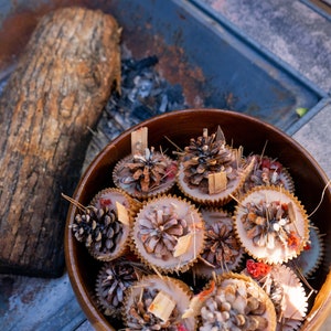 May include: A wooden bowl filled with pine cone candles. The candles are white with brown pine cones embedded in the wax. The bowl is sitting on a grey surface.