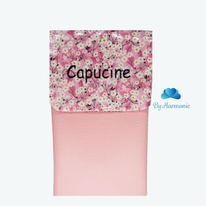 Puede incluir: Un estuche rosa con una solapa estampada con flores con la palabra "Capucine" en negro. La solapa tiene un fondo rosa con flores blancas y rosas. El estuche es de color rosa liso.