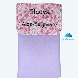 Puede incluir: Un estuche de color lavanda con una solapa estampada con flores. La solapa presenta el nombre "Gladys" y las palabras "Aide-Soignante" en texto negro. El estuche tiene una textura suave y un fondo claro.