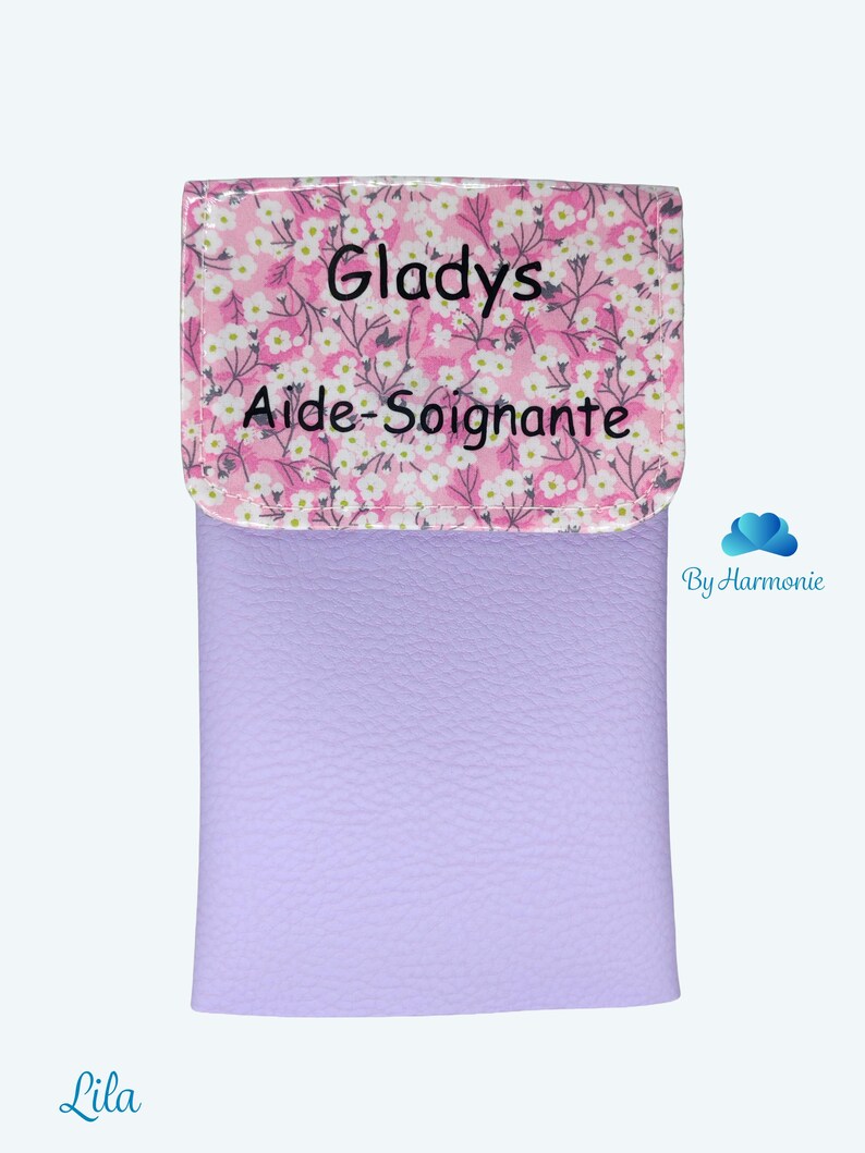 Puede incluir: Un estuche de color lavanda con una solapa estampada con flores. La solapa presenta el nombre "Gladys" y las palabras "Aide-Soignante" en texto negro. El estuche tiene una textura suave y un fondo claro.