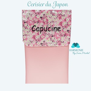 Puede incluir: Una bolsa rosa con una solapa estampada con flores blancas sobre un fondo rosa. La solapa tiene la palabra "Capucine" bordada en negro. El texto "Pochette Cerisier du Japon" est&aacute; encima de la solapa, y "Rose" en la parte inferior.