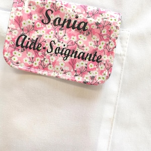 Puede incluir: Una etiqueta rectangular con un estampado floral rosa y el nombre "Sonia Aide-Soignante" en escritura negra. La etiqueta est&aacute; adherida a una prenda blanca, probablemente un uniforme o bata de laboratorio.