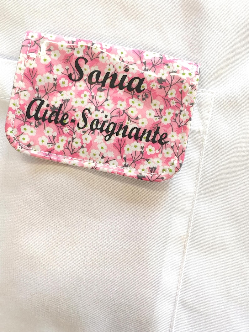 Puede incluir: Una etiqueta rectangular con un estampado floral rosa y el nombre "Sonia Aide-Soignante" en escritura negra. La etiqueta est&aacute; adherida a una prenda blanca, probablemente un uniforme o bata de laboratorio.