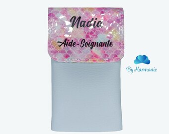 May include: A light blue wallet with a flap featuring a pink, white, and yellow scale pattern. The flap has the name "Nadia" and the words "Aide-Soignante" printed in black. The wallet is a light blue color.