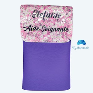 Puede incluir: Un estuche cil&iacute;ndrico violeta con una superficie texturizada. La solapa superior presenta un dise&ntilde;o floral rosa con el nombre "Stefanie" y las palabras "Aide-Soignante" en escritura negra. Un accesorio pr&aacute;ctico.