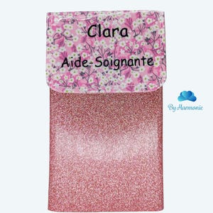 Puede incluir: Un estuche rosa brillante con una solapa estampada floral. La solapa tiene el nombre "Clara" y las palabras "Aide-Soignante" impresas. La parte inferior del estuche tiene las palabras "Rose paillette" impresas.