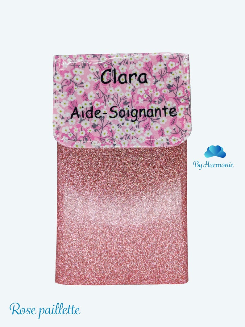 Puede incluir: Un estuche rosa brillante con una solapa estampada floral. La solapa tiene el nombre "Clara" y las palabras "Aide-Soignante" impresas. La parte inferior del estuche tiene las palabras "Rose paillette" impresas.
