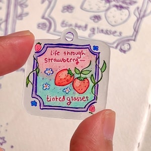 Puede incluir: Un amuleto de plástico transparente con un diseño dibujado a mano de dos fresas, enredaderas y flores. El amuleto tiene un borde morado y el texto "life through strawberry - tinted glasses".