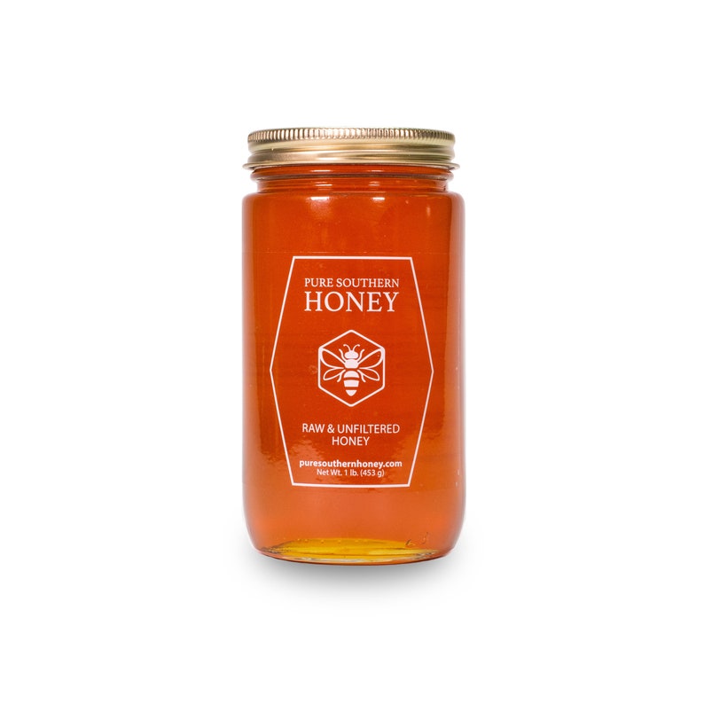 Puede incluir: Un tarro de vidrio de miel con una tapa dorada. La etiqueta dice "Pure Southern Honey" con una ilustraci&oacute;n de abeja y el texto "Miel cruda y sin filtrar" y "puresouthernhoney.com". El tarro contiene 1 libra (453 gramos) de miel.