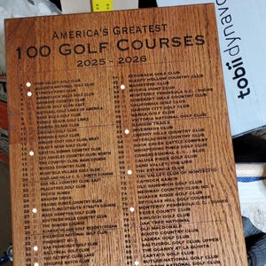 May include: Wooden plaque listing "America's Greatest 100 Golf Courses" for 2025-2026. The plaque is brown with black text, featuring a list of golf clubs numbered 1-100.