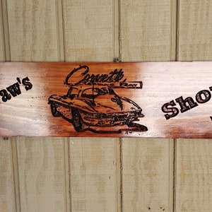 Op de afbeelding: Een houten bord met de tekst "Paw Paw's Corvette Shop" en een zwart-wit afbeelding van een Corvette-auto. Het bord heeft aan beide kanten geblokte vlaggen.