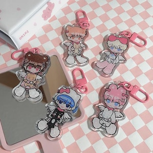 May include: Five clear acrylic keychains with pink lobster clasps. Each keychain features a cartoon angel character with wings, holding a small plush toy. The keychains are on a pink and white checkered surface.