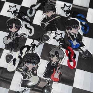 May include: Keychains featuring anime-style illustrations of musicians playing guitars. Each keychain has a unique guitar color, including white, blue, and red. The keychains are attached to black and white chain links and star-shaped charms.