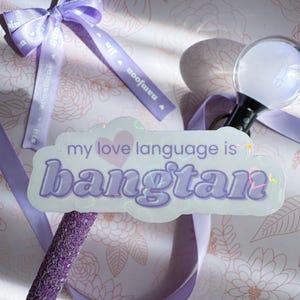 May include: A light purple sticker with the text "my love language is bangtan" is the focal point of this image. A matching purple ribbon and a light stick are also visible. The background features a floral pattern.
