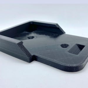 K&ouml;nnte beinhalten: Ein schwarzer, 3D-gedruckter Telefonst&auml;nder aus Kunststoff mit rechteckiger Basis und einer erh&ouml;hten Plattform. Der St&auml;nder hat ein kleines Loch und einen Schlitz oben.