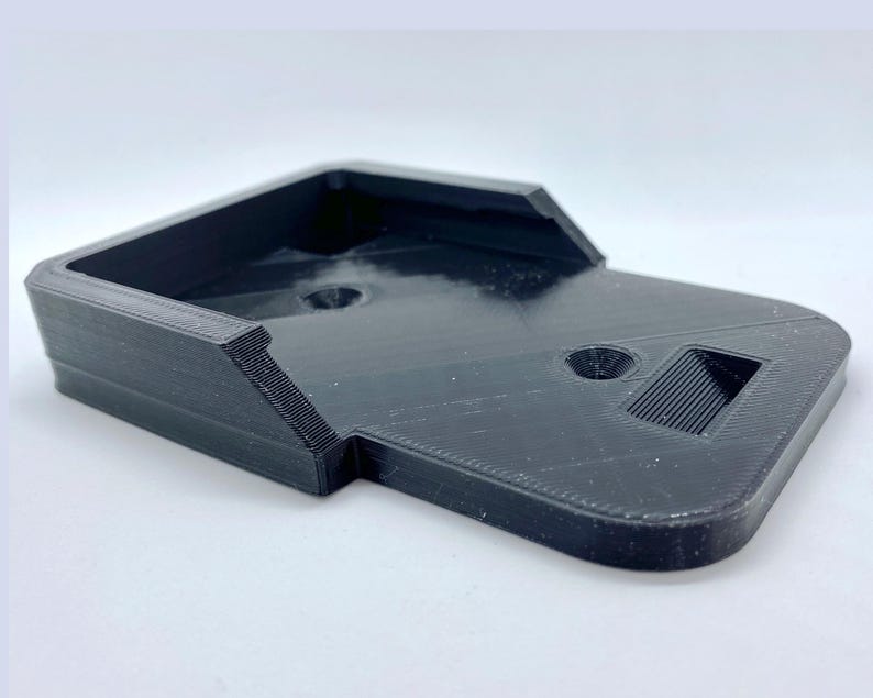K&ouml;nnte beinhalten: Ein schwarzer, 3D-gedruckter Telefonst&auml;nder aus Kunststoff mit rechteckiger Basis und einer erh&ouml;hten Plattform. Der St&auml;nder hat ein kleines Loch und einen Schlitz oben.