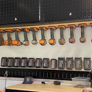 K&ouml;nnte beinhalten: Ein Wand-Aufbewahrungssystem mit mehreren orangefarbenen und schwarzen Elektrowerkzeugen, die daran h&auml;ngen. Unter dem Regal befinden sich mehrere schwarze und orangefarbene Batterien, die auf einem Regal aufgereiht sind.