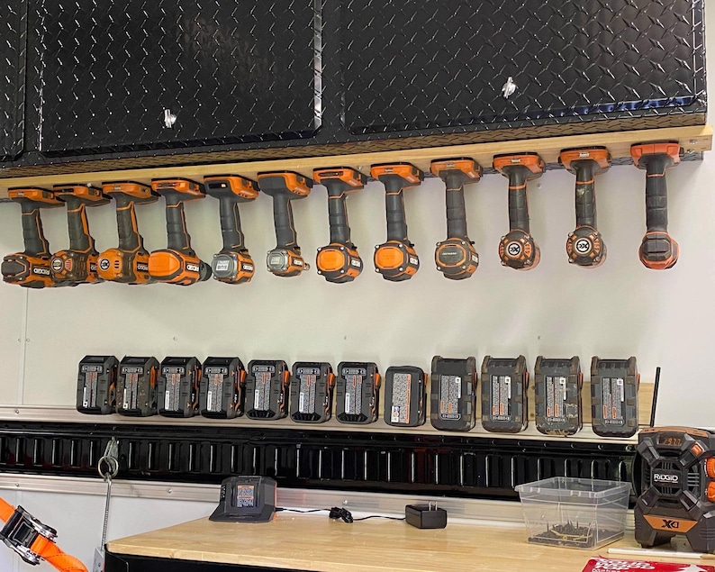 K&ouml;nnte beinhalten: Ein Wand-Aufbewahrungssystem mit mehreren orangefarbenen und schwarzen Elektrowerkzeugen, die daran h&auml;ngen. Unter dem Regal befinden sich mehrere schwarze und orangefarbene Batterien, die auf einem Regal aufgereiht sind.